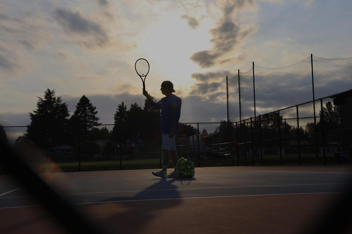 Girls varsity tennis coach Mr. David Lane holds his racquet to the sky under the sunlit clouds following practice.